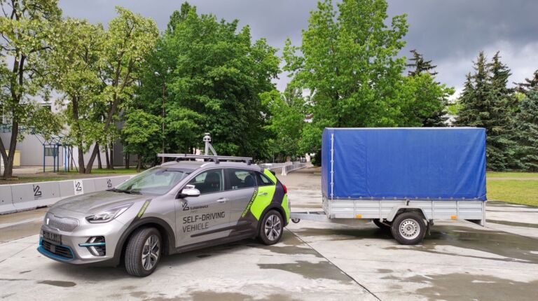 Stojący na betonowym placu szary samochód z logo Łukasiewicz z podpiętą naczepą